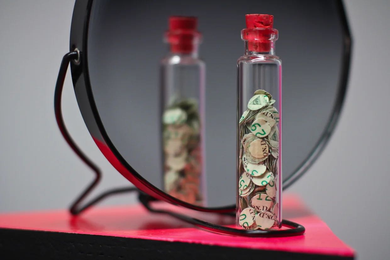 A glass vial filled with circular punch-outs from American one-dollar bills, reflected in a mirror.